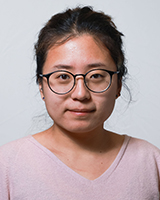 Une femme asiatique aux cheveux noirs, attachés, porte des lunettes à monture noire épaisse et un chandail rose pâle à encolure en V. L’arrière-plan est gris pâle.