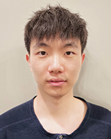 Un homme asiatique dans la vingtaine, aux cheveux crépus et noirs, de longueur moyenne. Son expression est neutre et il porte un chandail bleu marine de marque Henley. Il a été photographié devant un mur blanc.