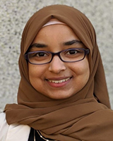 Une femme aux yeux brun foncé sourit. Elle porte des lunettes à monture noire, un hidjab brun, un chandail noir, un veston blanc et deux colliers dorés. L’arrière-plan blanc et gris est flou.