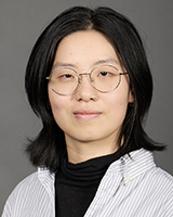 Une femme asiatique aux cheveux noirs mi-longs, portant des lunettes à monture argentée et un anneau de la même couleur dans le nez. Elle porte un chandail à col roulé noir ainsi qu’une chemise blanche à fines rayures verticales grises. L’arrière-plan est gris foncé.