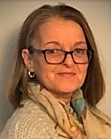 Une femme au teint pâle et aux cheveux brun clair sourit. Elle porte des lunettes à monture noire épaisse et de petits anneaux aux oreilles, ainsi qu’une veste de laine blanche et un foulard beige à motifs bleus, bruns et jaunes. L’arrière-plan est gris pâle.